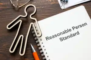 A figurine of a human rests on a wood desk with a book. The title reads “Reasonable Person Standard.”