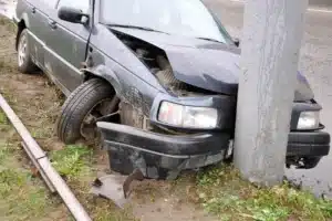 A dark, four-door car, with a crumpled front-end. A result of a single-vehicle accident of hitting a light pole, which it rests against still.