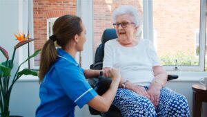 Senior Woman Sitting In Chair In Residents Lounge And Talking With Nurse In Retirement Home